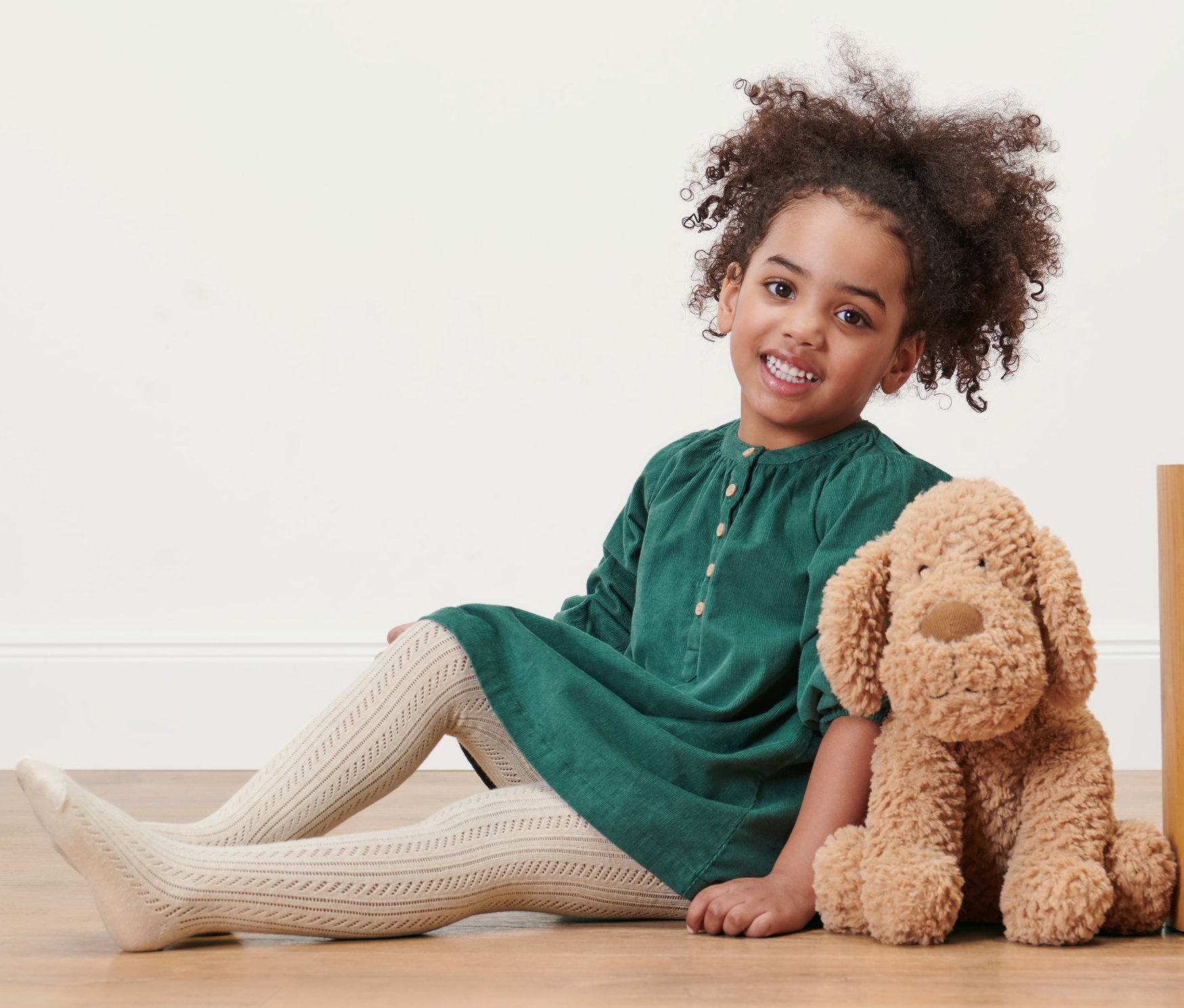 Usmívající se holčička sedí na podlaze v zelených šatech a má na sobě dětské punčocháče, 2 páry. Vedle ní sedí plyšový pes.