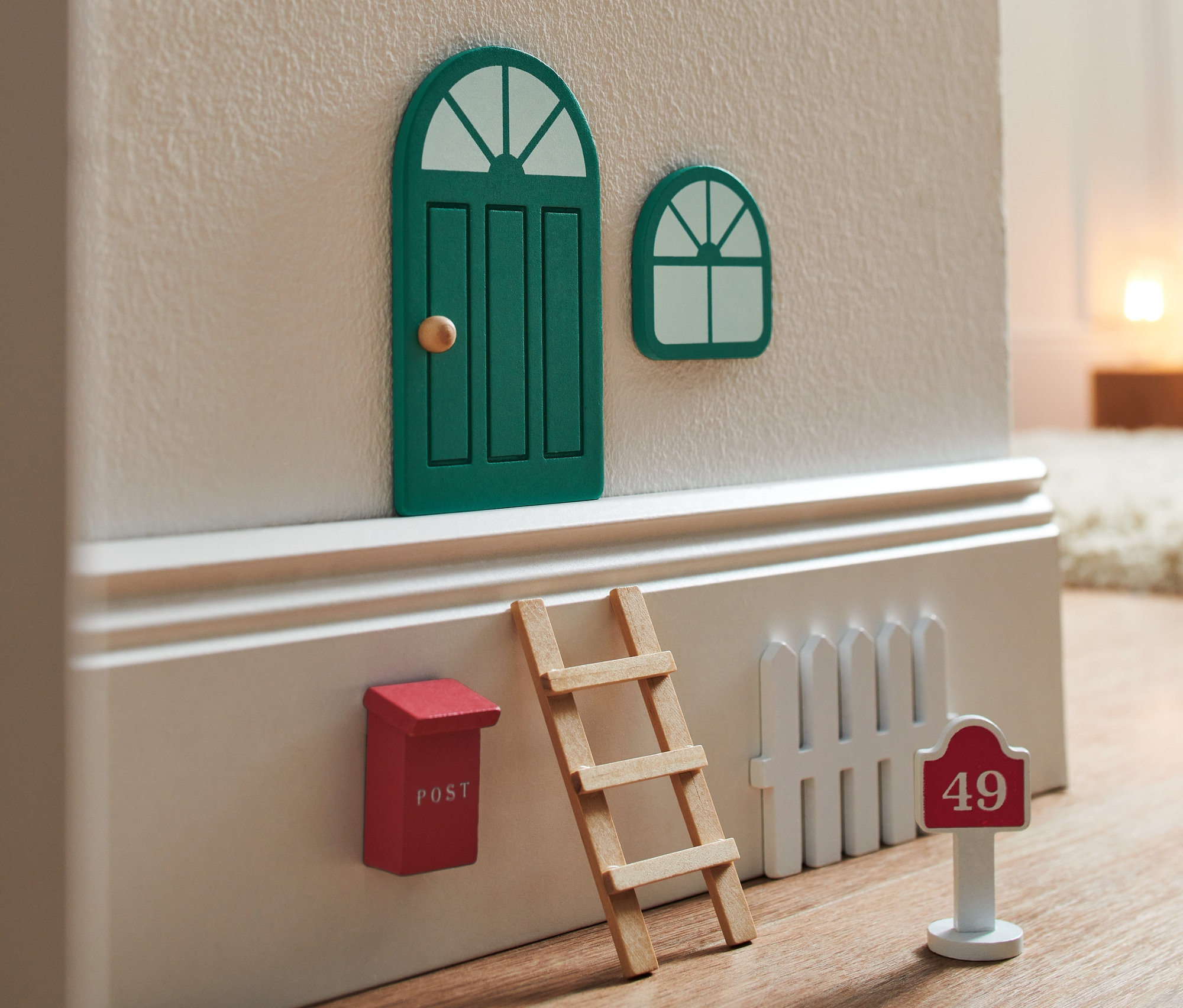 Zavřené zelené dveře a okno Dvířka pro skřítka s doplňky, červená poštovní schránka, žebřík a bílý plot s číslem domu 49.