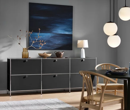 Černá odkládací skříňka CN3 se 6 výklopnými dvířky, bílá stolní lampa s textilním stínidlem a závěsné svítidlo se skleněnými koulemi.