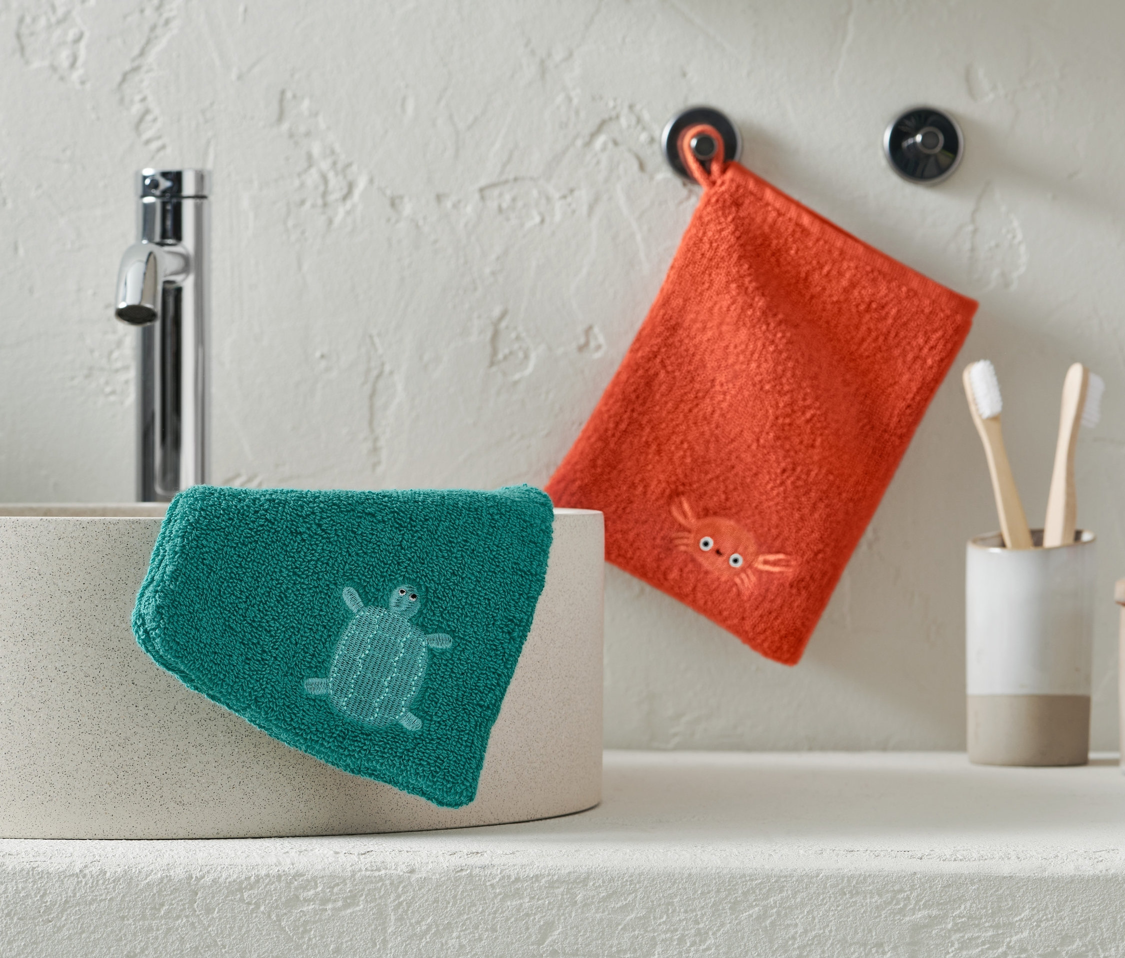 Zelená žínka s želvou leží na umyvadle, oranžová žínka s krabem visí na zdi.