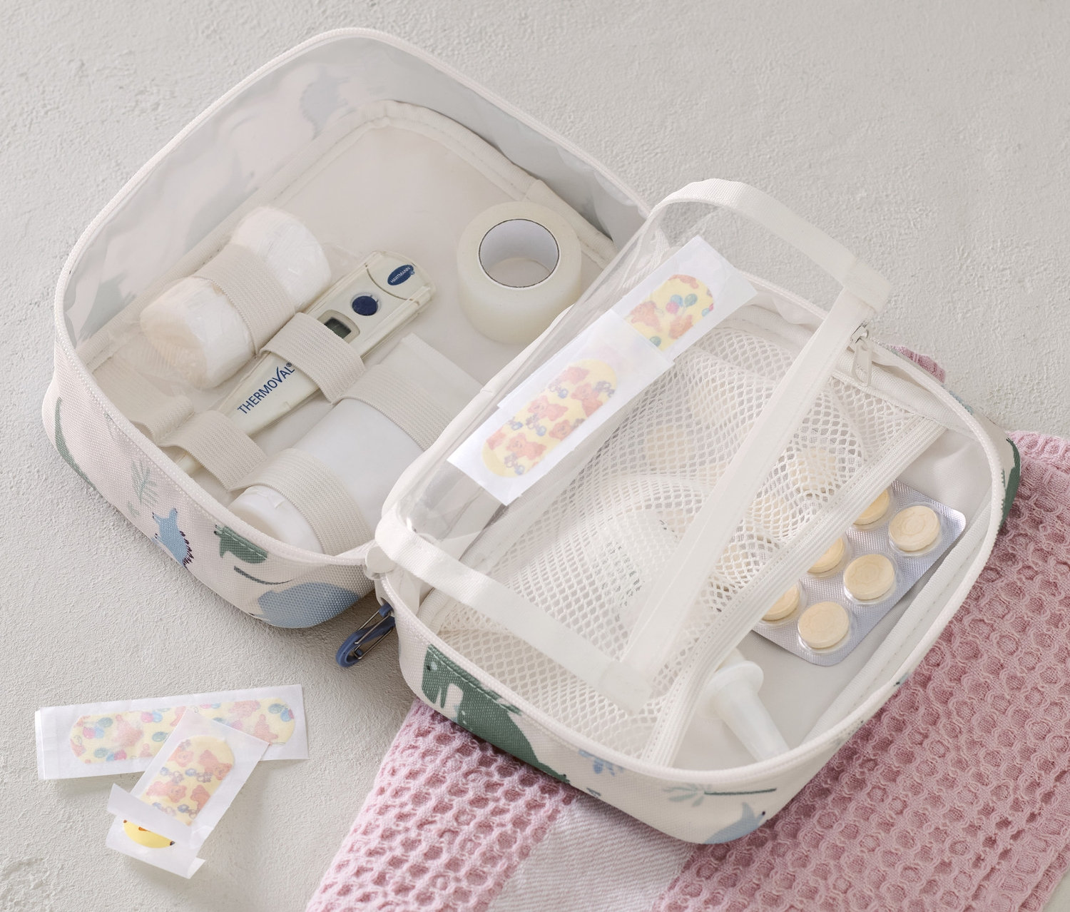 Otevřená taštička na léky s obsahem a náplastmi leží na růžovém ručníku.