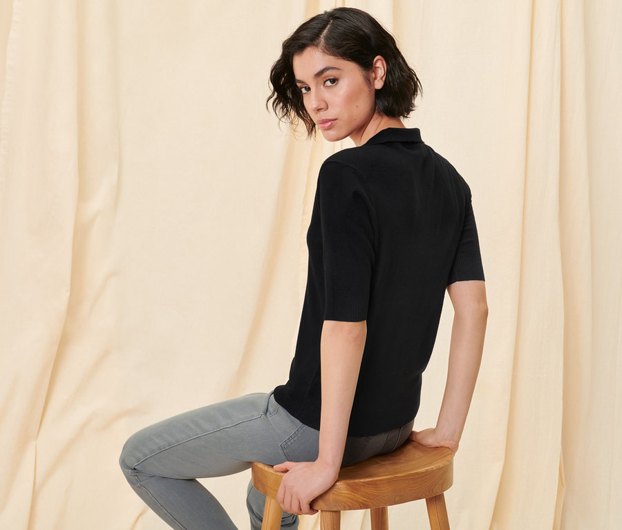 Žena sedí na dřevěné stoličce a má na sobě černé polo z jemného úpletu a světle šedé tregíny.
