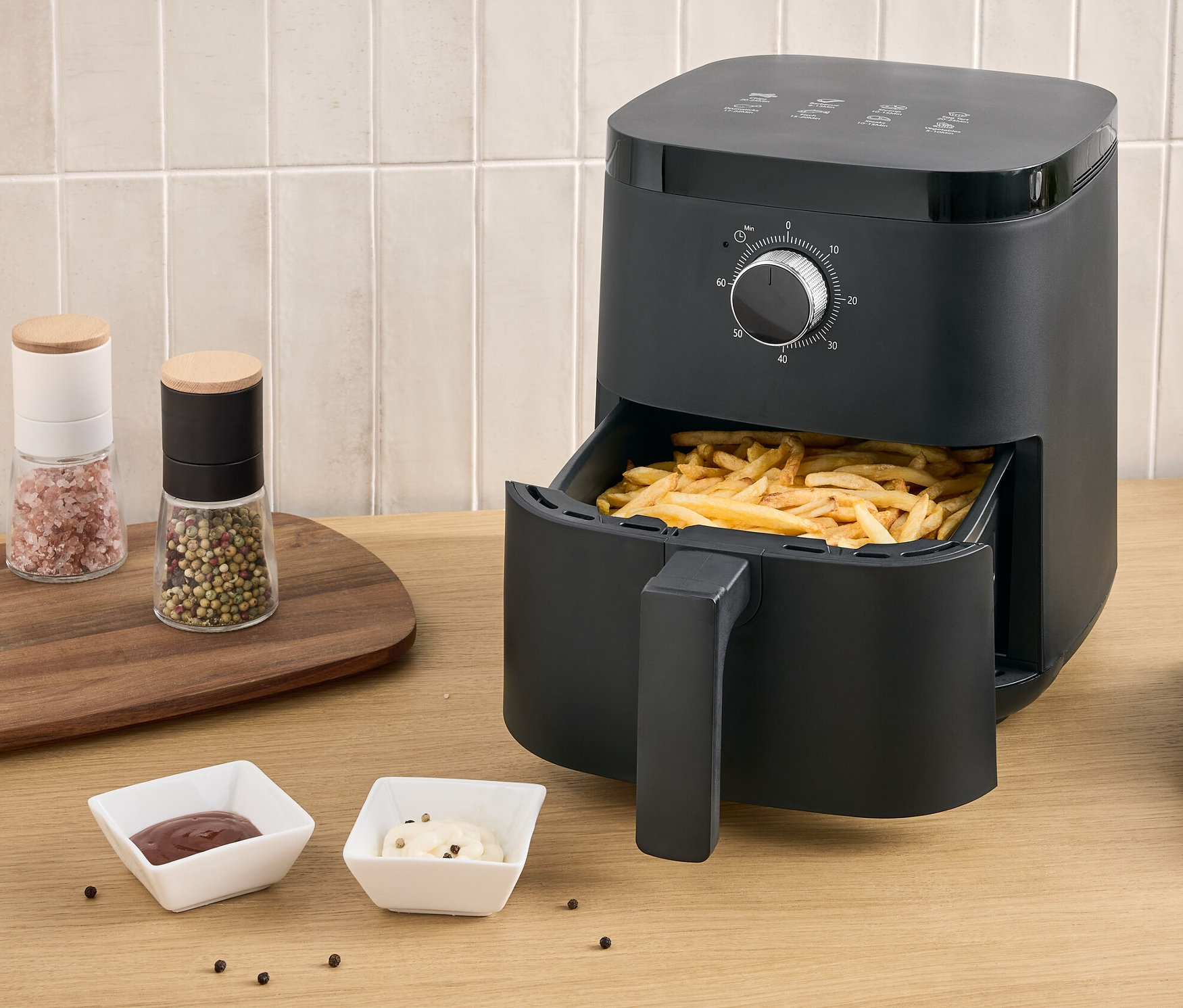 Černá horkovzdušná fritéza s hranolky ve výsuvné zásuvce stojí na stole vedle talíře s hranolky a dvou omáček.