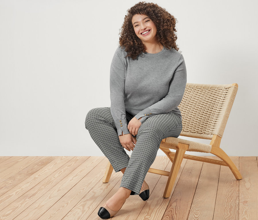 Usmívající se žena sedí na židli, má na sobě šedý melírovaný svetr a vzorované strečové kalhoty.