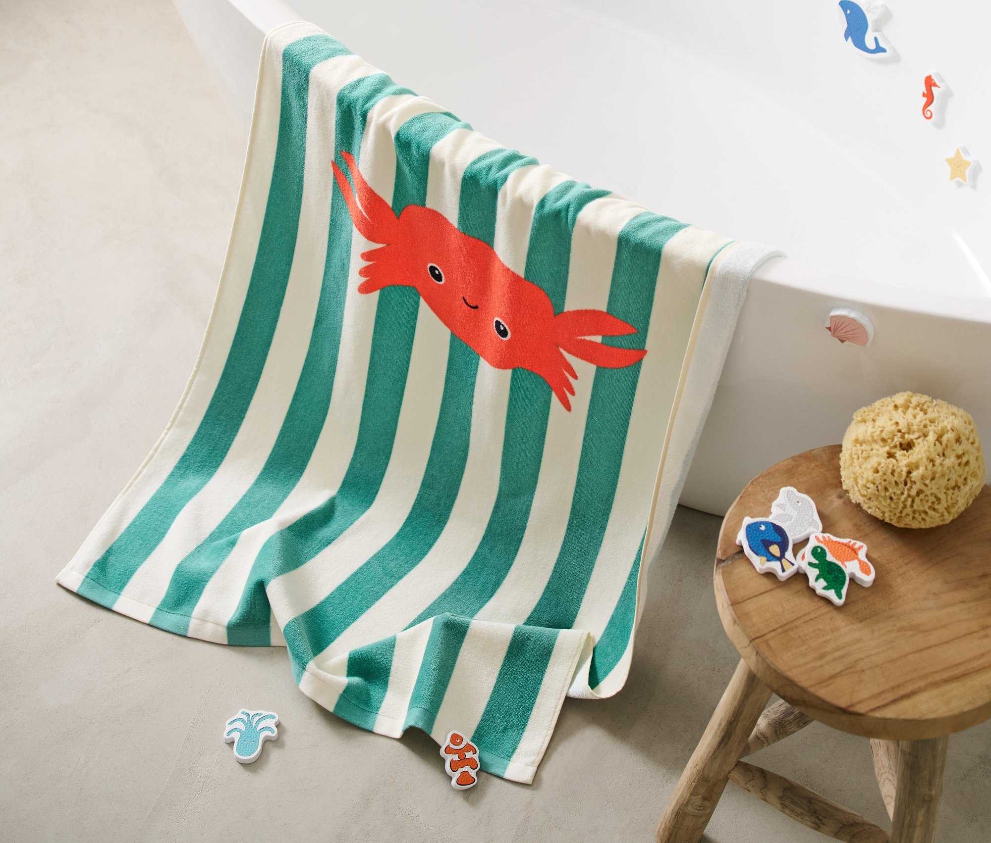 Pruhovaná osuška s motivem kraba leží okraji vany, vedle vany stojí dřevěná stolička s mořskou houbou.