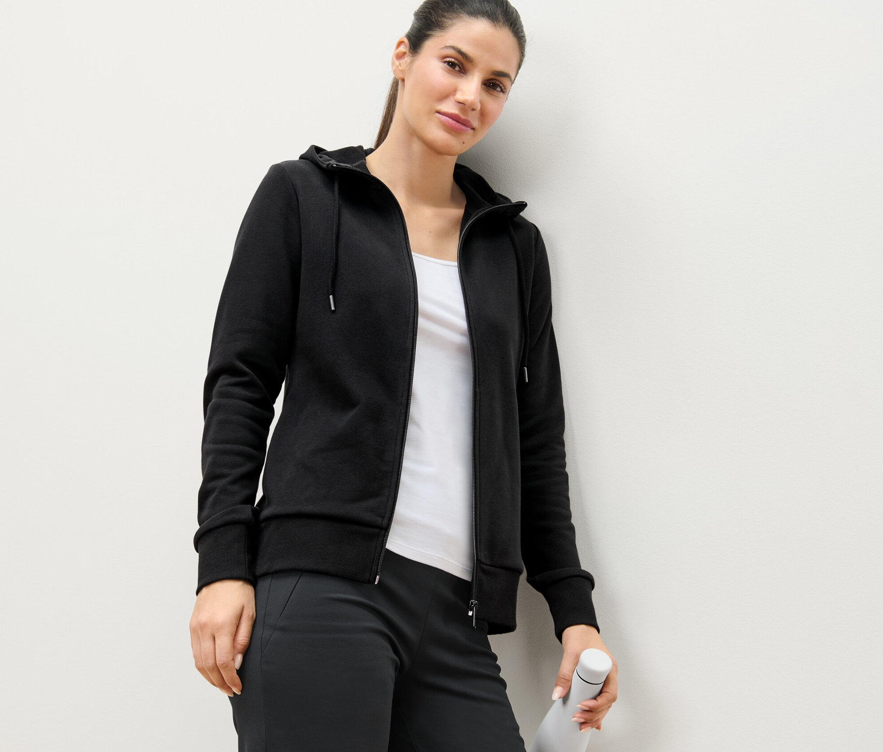 Žena opřená o zeď má na sobě černou mikinu a drží bílou láhev s vodou.