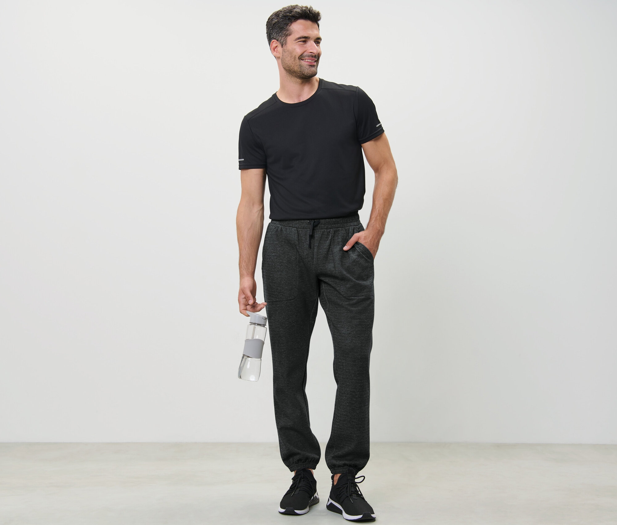 Muž v černém tričku drží láhev s vodou a má na sobě teplákové kalhoty.