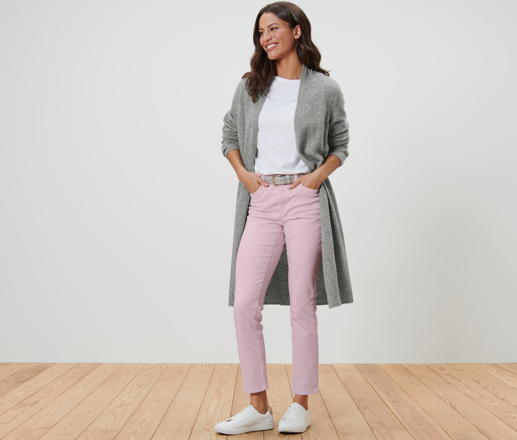 Žena v otevřeném dlouhém kardiganu, bílém tričku, růžových kalhotách a bílých teniskách stojí na dřevěné podlaze.