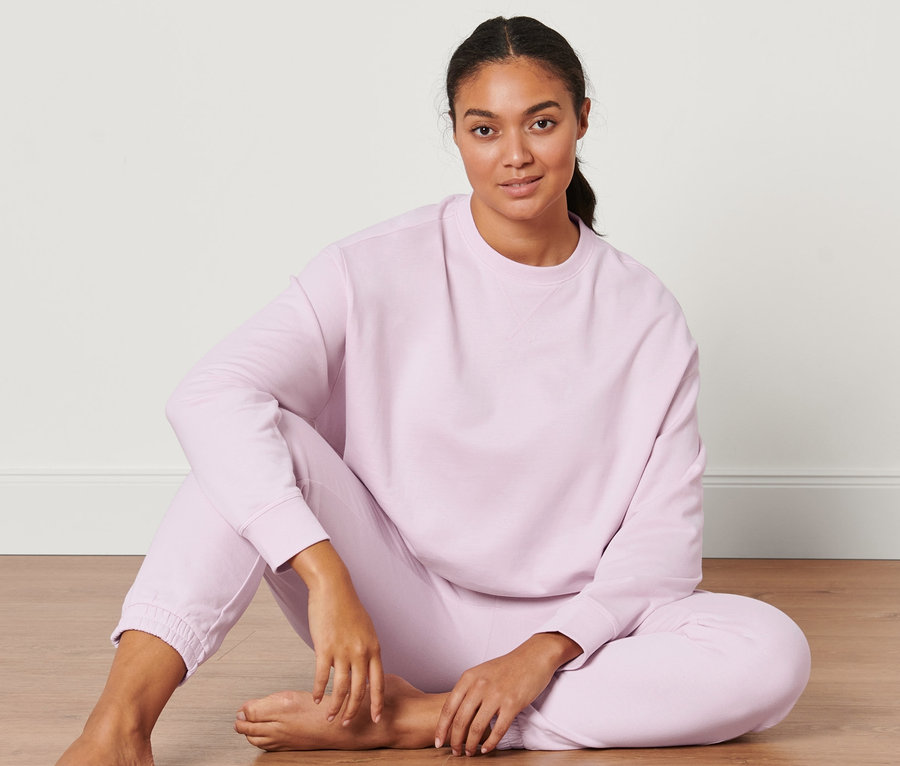Žena sedí na podlaze a má na sobě světle růžovou oversized mikinu na jógu.