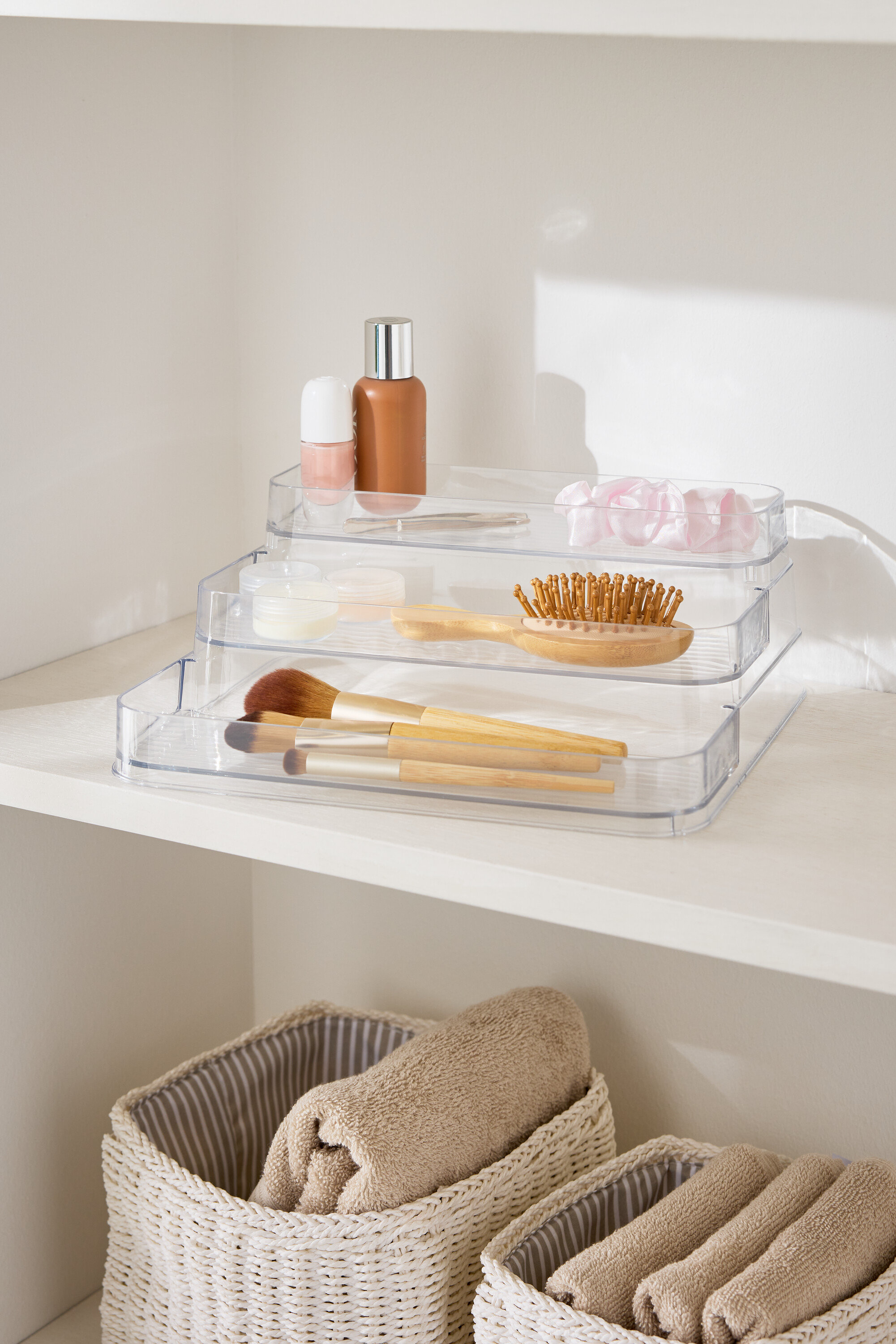 Třípatrový průhledný organizér na líčení na bílé polici vedle košíků s ručníky.