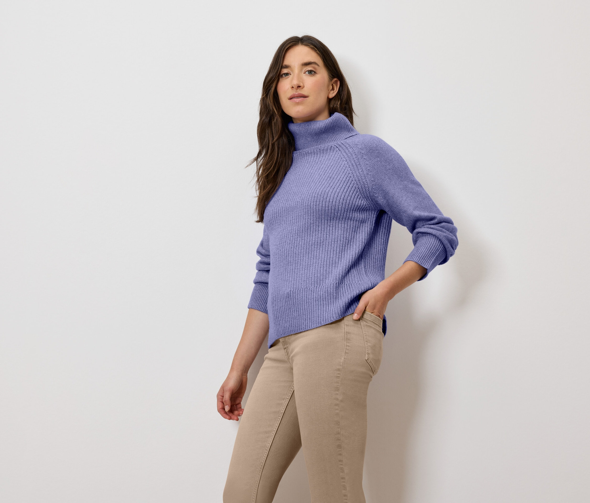 Žena pózuje v lila roláku a béžových kalhotách.