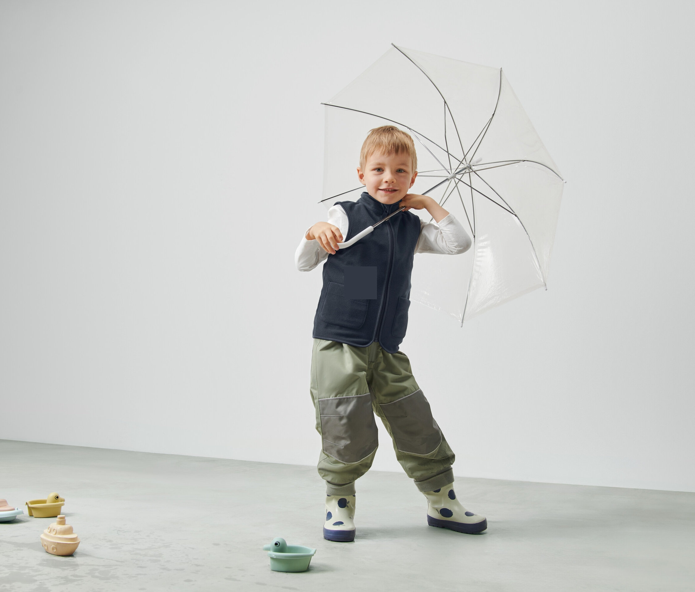 Chlapec drží průhledný deštník, má na sobě vestu a kalhoty a stojí v místnosti.