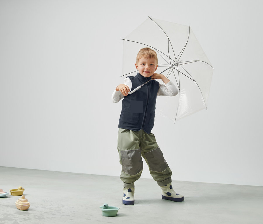 Chlapec drží průhledný deštník, má na sobě vestu a kalhoty a stojí v místnosti.