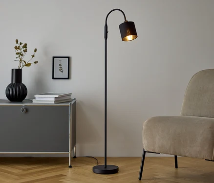 Černá stojací lampa stojí vedle šedé komody, na které je váza a knihy. Vedle komody stojí béžové křeslo.
