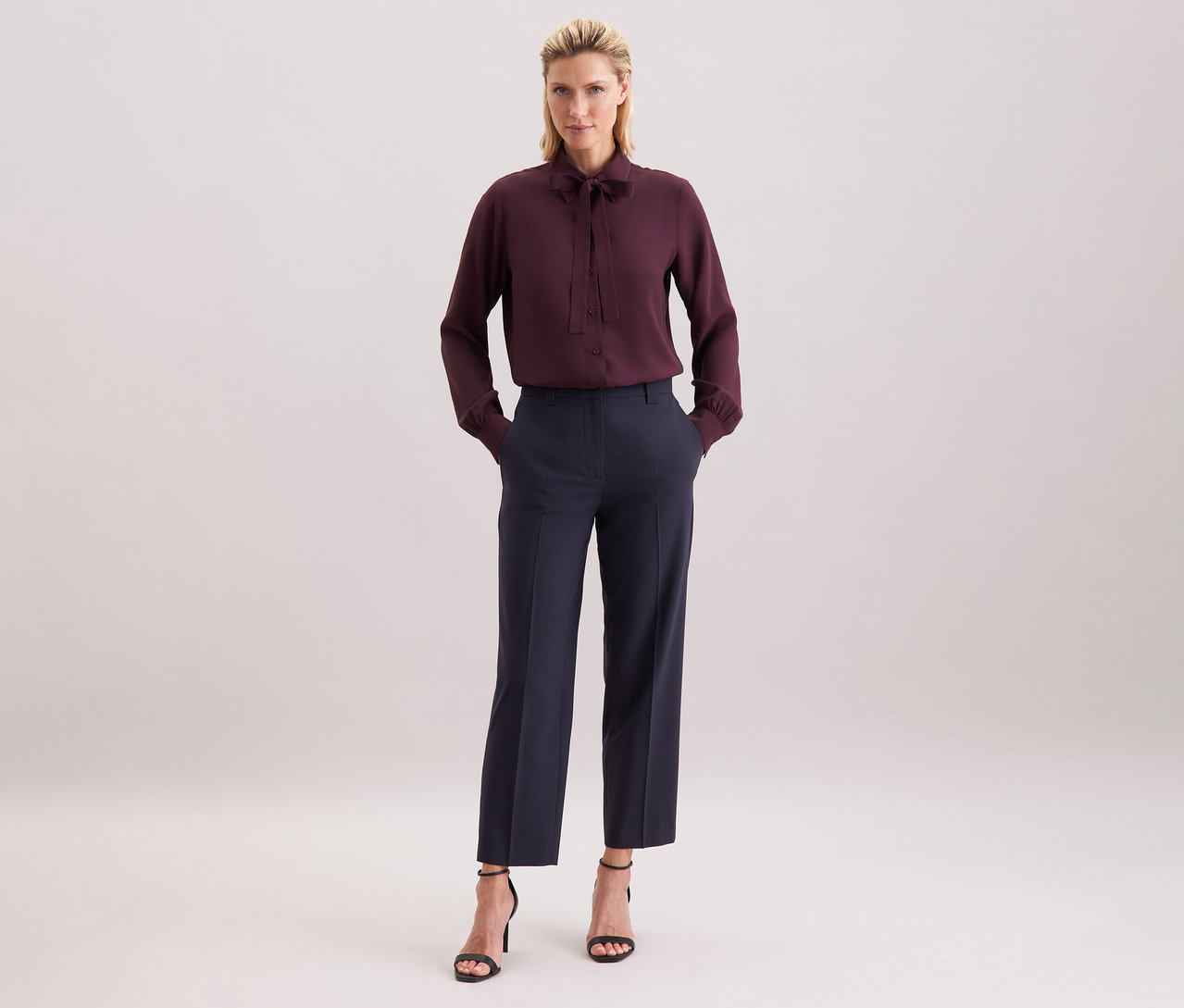 Žena má na sobě vínovou halenku s vázačkou Seidensticker z viskózy, tmavé kalhoty a černé sandály.