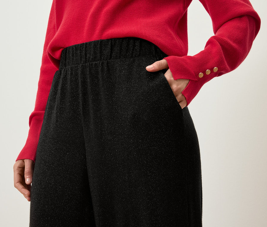 Částečný pohled na ženu v červeném svetru s rukou v kapse černých třpytivých kalhot.