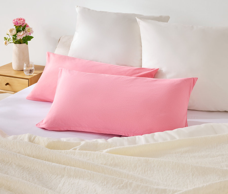 Na posteli leží dva růžové a dva bílé polštáře. Na nočním stolku je váza s květinami.