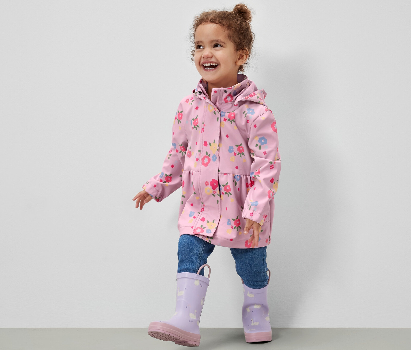 Usmívající se holčička v dětské softshellové bundě s květinovým vzorem, džínách a fialových holínkách kráčí po šedé podlaze před bílou zdí.