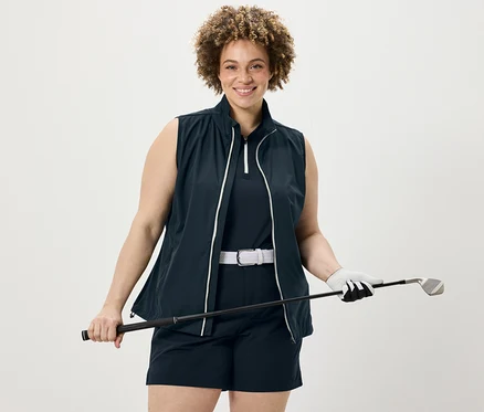 Žena drží golfovou hůl, má na sobě tmavě modrý funkční top, softshellové funkční šortky a softshellovou vestu.