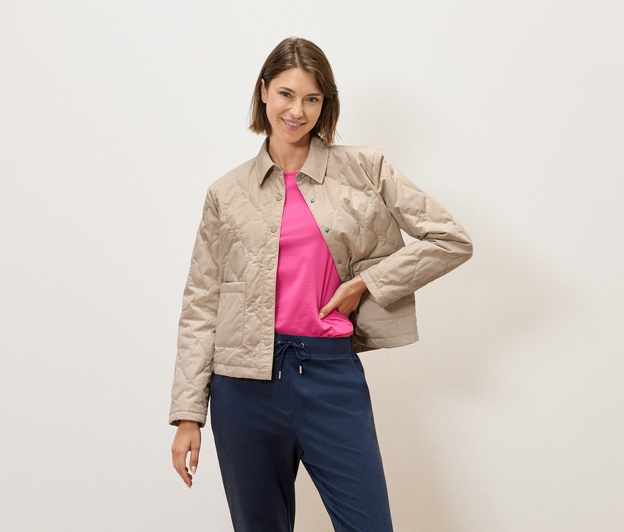 Žena s krátkými hnědými vlasy pózuje v béžové prošívané bundě, růžovém tričku a modrých kalhotách na pozadí krémové zdi.