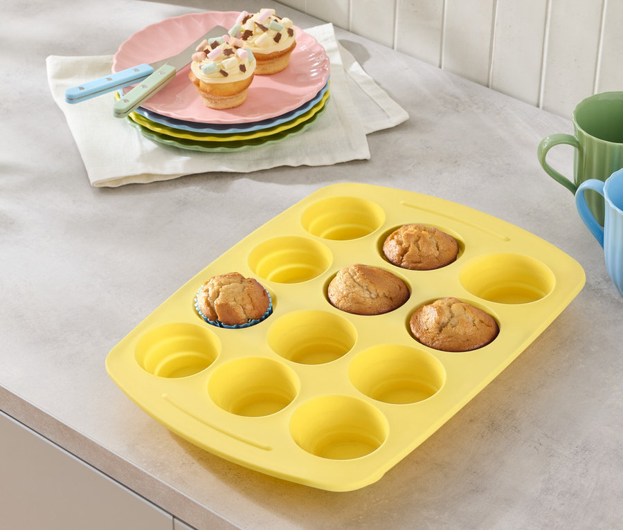 Žlutá forma na muffiny s několika muffiny, talíře s muffiny a sklenice na pracovní desce.