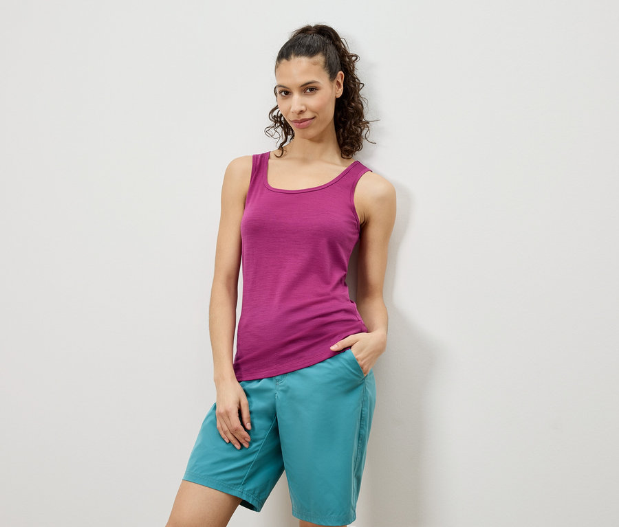 Žena pózuje v růžovém tílku a tyrkysových šortkách před bílou stěnou.