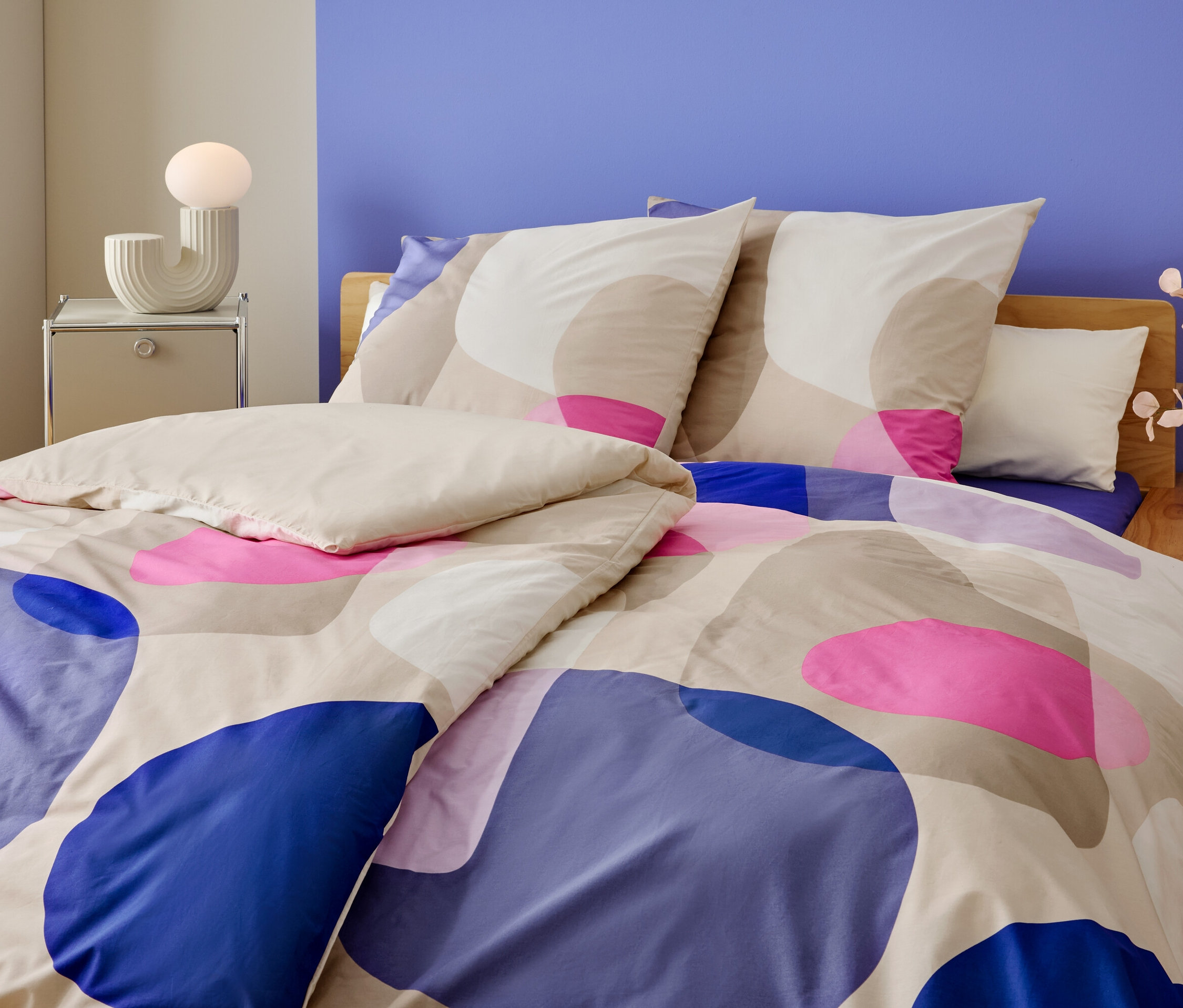 Detailní záběr postele s geometrickým vzorem v modré, růžové a béžové barvě. Na nočním stolku stojí lampa a váza s květinami.