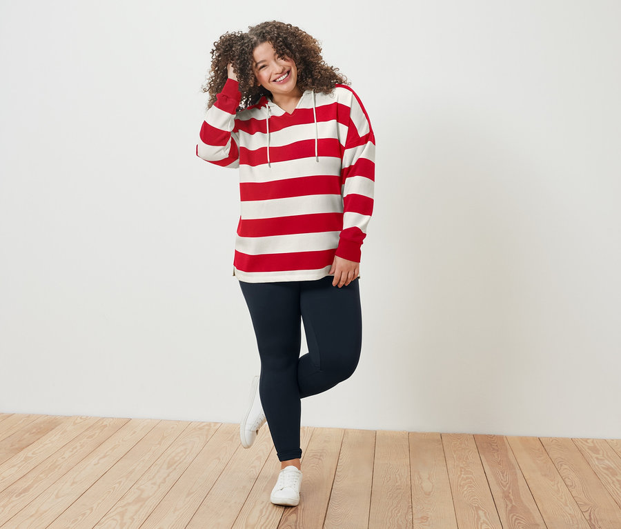 Žena s kudrnatými vlasy v červeno-bílém pruhovaném topu a modrých legínách stojí na dřevěné podlaze.