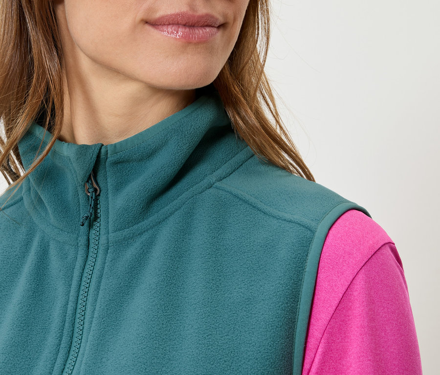 Detail ženy v petrolejové fleecové vestě přes růžový top.