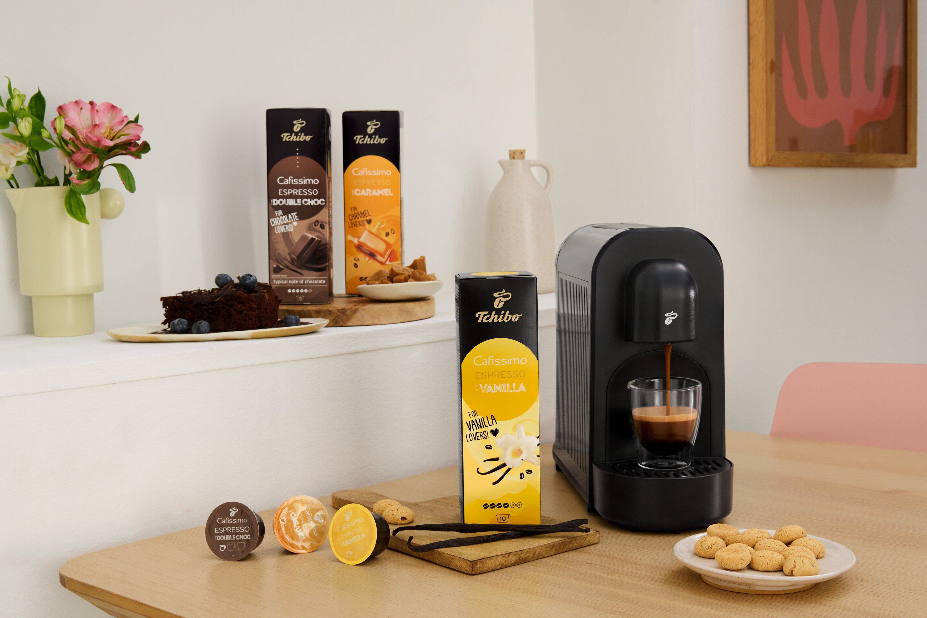 Na stole stojí kávovar, tři balení kapslí Cafissimo (Double Choc, Vanilka, Karamel), sušenky a kousek čokoládového dortu.