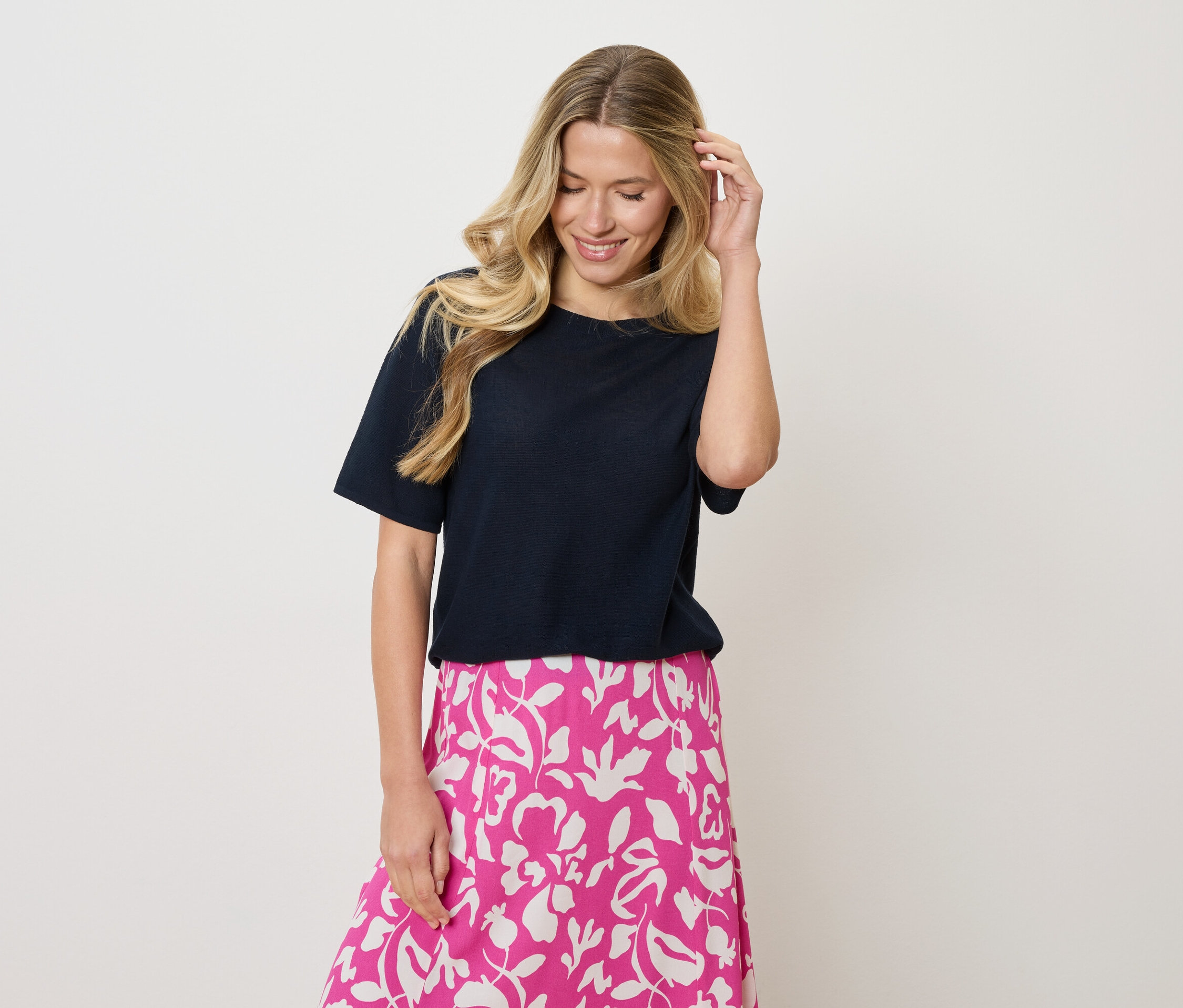 Žena s blond vlnitými vlasy nosí tmavě modré tričko a růžovo-bílou sukni s květinovým vzorem, dotýká se vlasů jednou rukou.