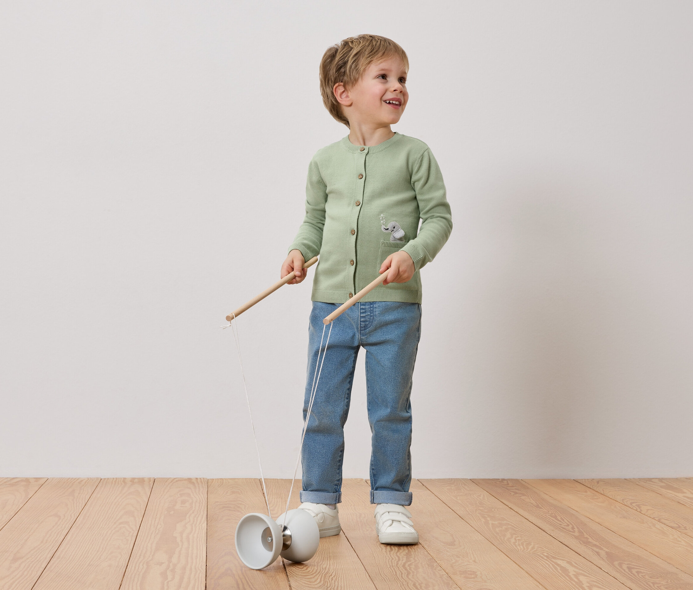 Usmívající se chlapec s blond vlasy si hraje s diabolem. Má na sobě zelený kardigan se sloním motivem, džíny a bílé tenisky.