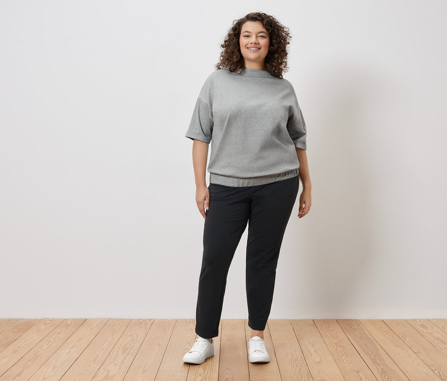 Žena s kudrnatými vlasy stojí před bílou zdí a má na sobě šedou mikinu, černé kalhoty a bílé tenisky.