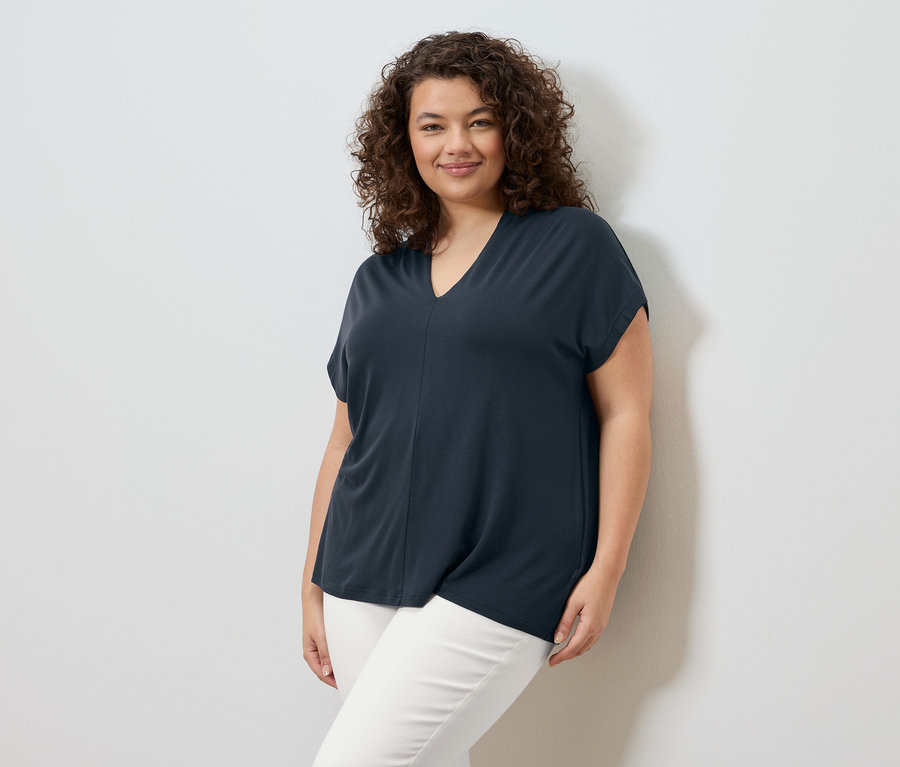 Žena s kudrnatými vlasy stojí před bílou zdí a má na sobě tmavě modré tričko a bílé kalhoty.