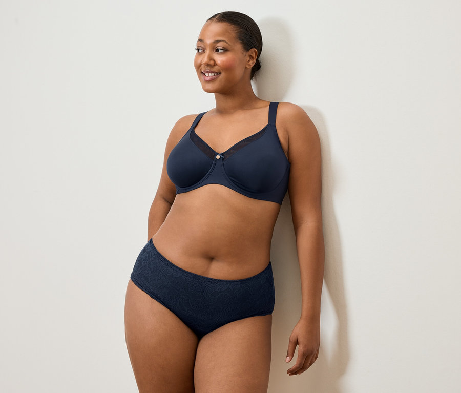 Žena v tmavě modré podprsence a kalhotkách stojí u bílé zdi a usmívá se.