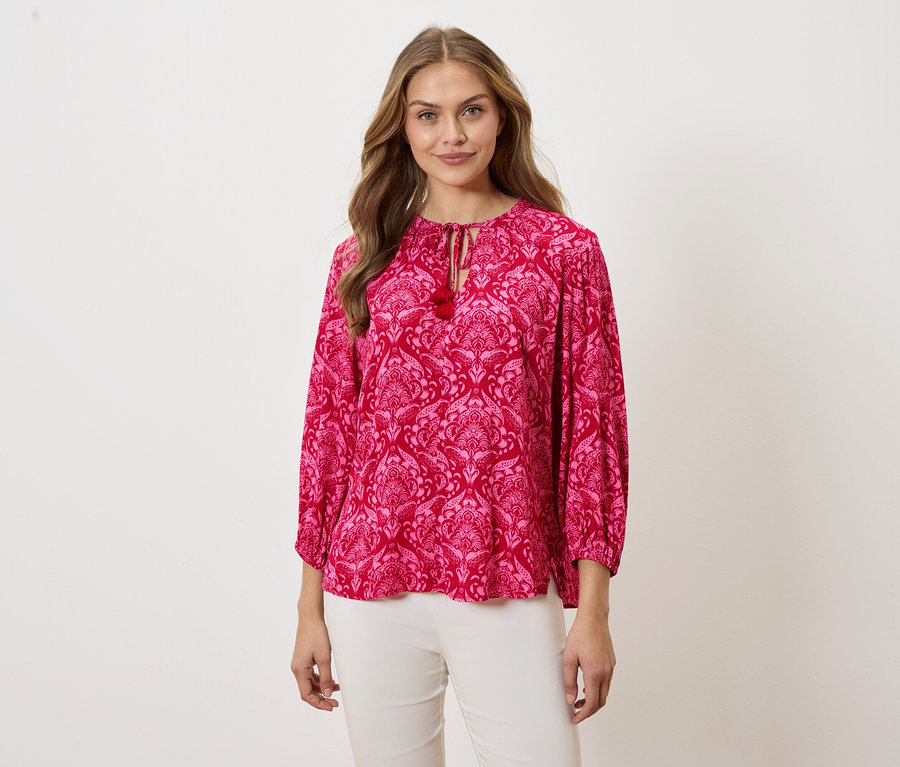 Žena má na sobě růžovou blůzu se vzorem a bílé kalhoty.