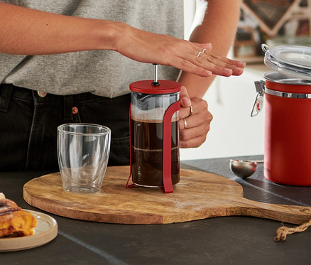 Usměvavá žena v brýlích stlačuje píst červené konvičky french press 300 ml na dřevěném prkénku, vedle jsou skořicoví šneci a prázdná sklenice.
