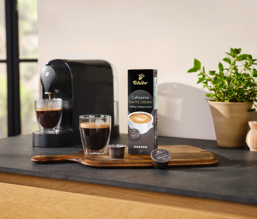 Kávovar připravuje kávu do sklenice vedle balení Cafissimo Caffè Crema silná – 10 kapslí.