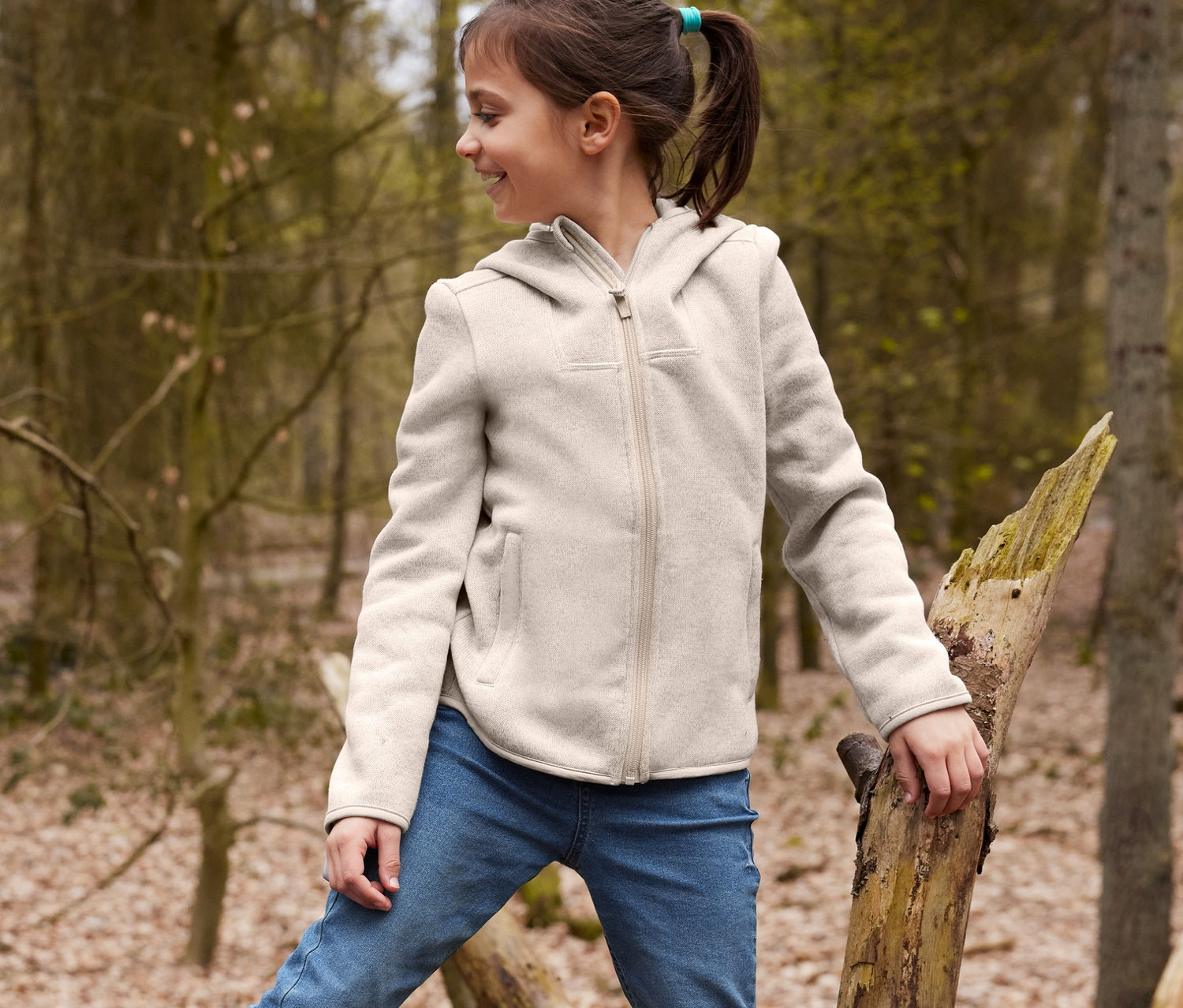 Chlapec sedí na spadlém stromě v žlutých holínkách z recyklovatelného materiálu a hnědé oboustranné čepici, má na sobě džíny a bundu z pleteného fleecu. Dívka stojí na stromě v džínách a bundě z pleteného fleecu.
