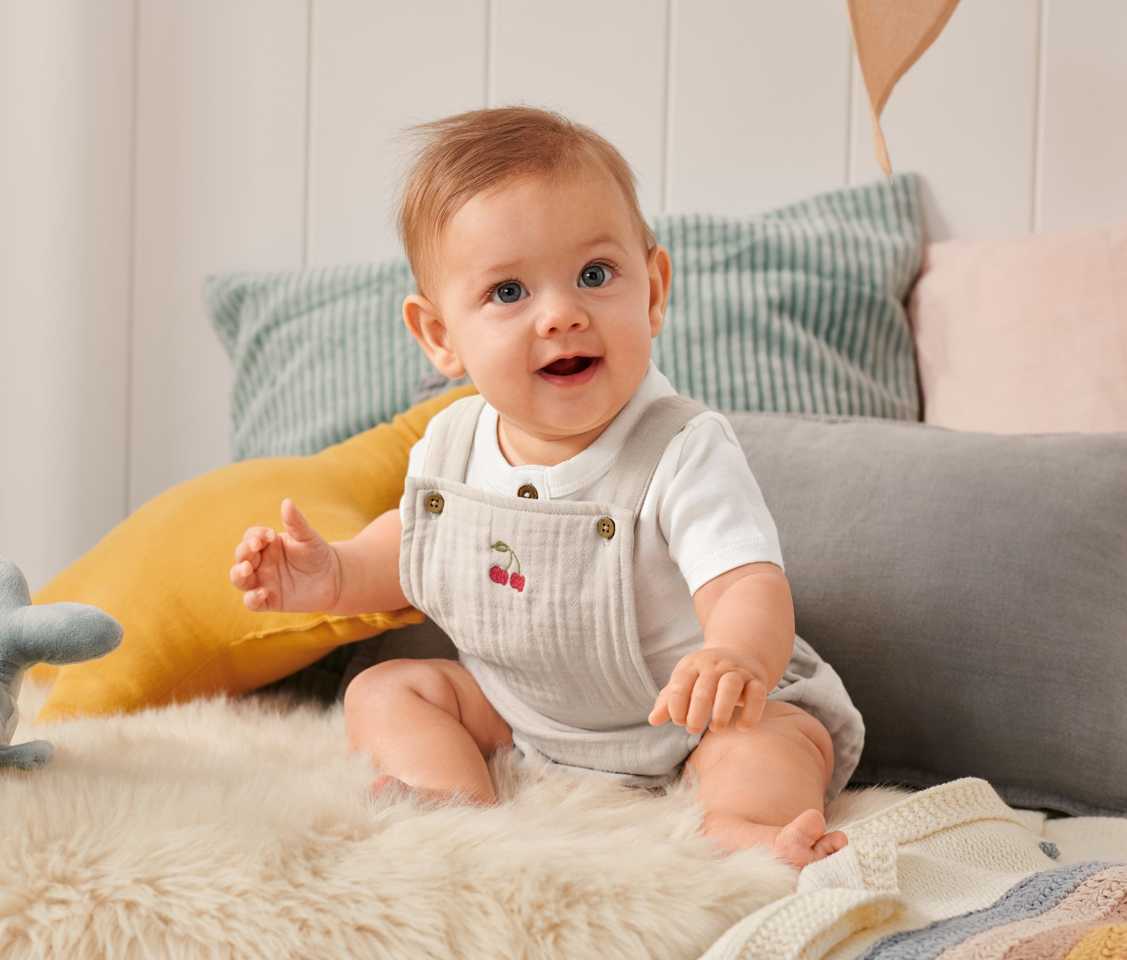 Usmívající se dítě sedí na kožešině a má na sobě krátké dupačky s výšivkou třešní.