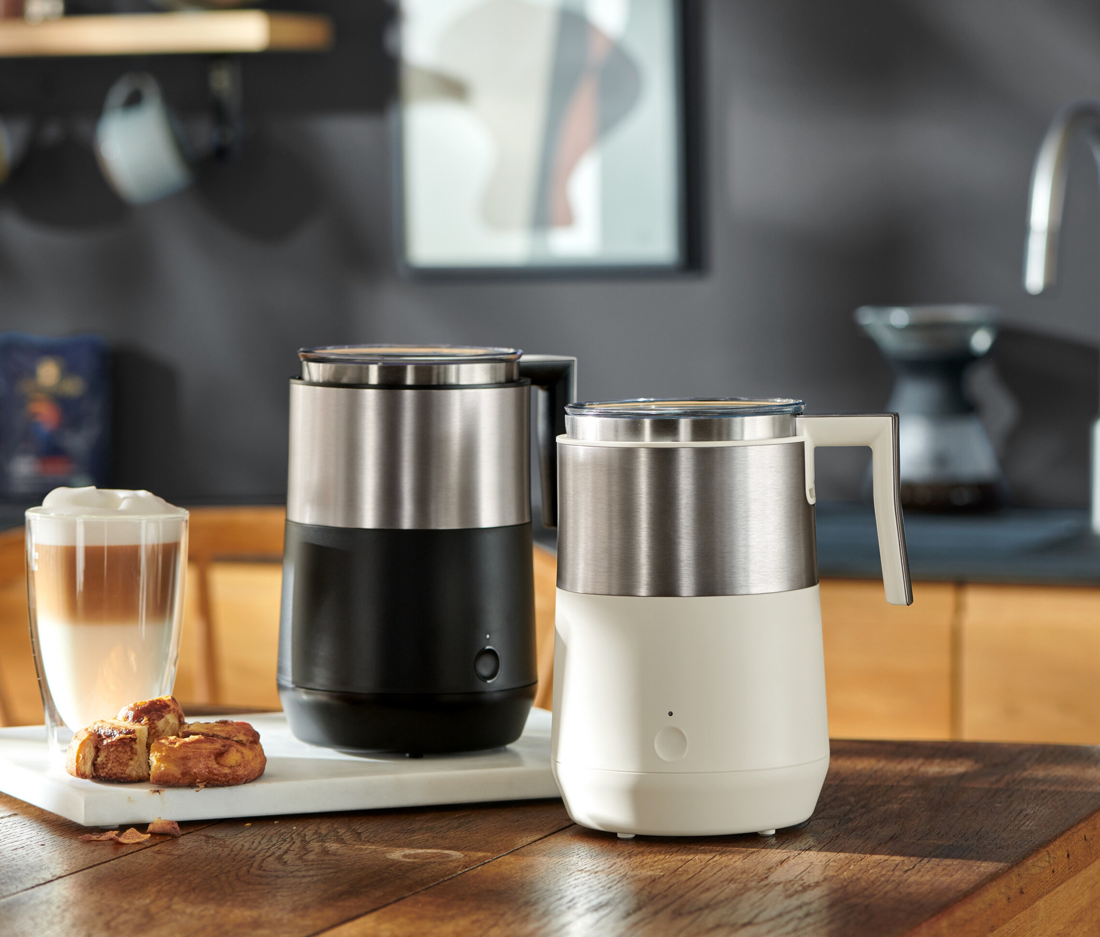 Dva indukční napěňovače mléka, jeden černý a jeden krémově bílý, stojí na dřevěném stole vedle sklenice latté a pečiva.