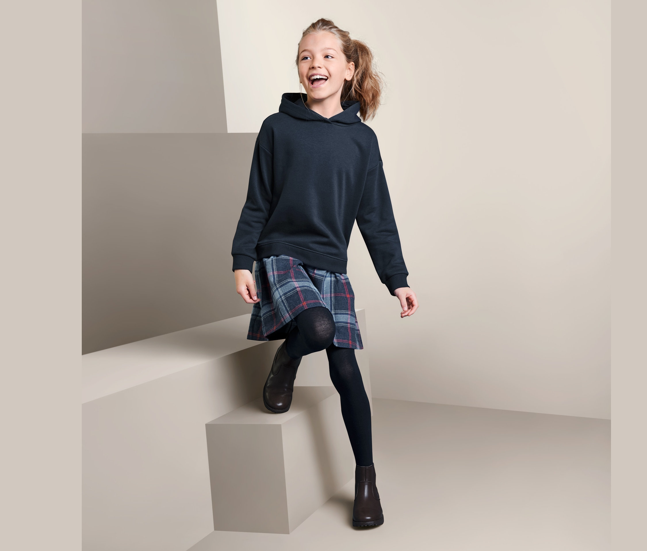 Usmívající se dívka sedí na schodech a má na sobě tmavě modré šaty z teplákoviny a kostkovanou sukni.