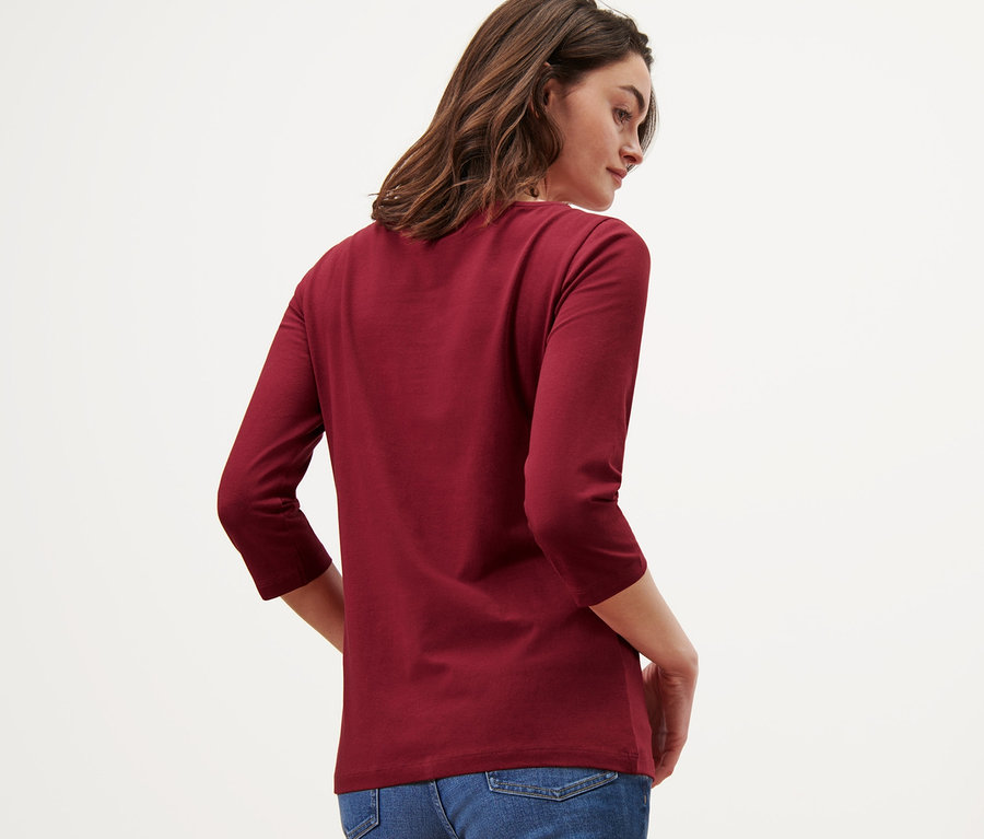 Žena pózuje zády, má na sobě vínově červené triko s 3/4 rukávem a džíny.