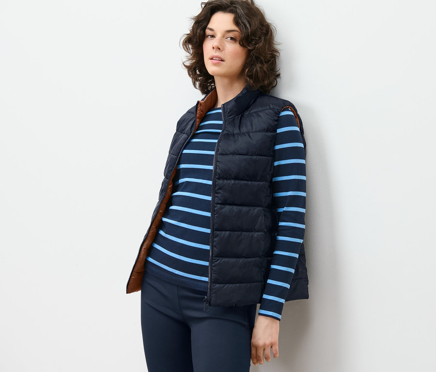 Žena opřená o zeď má na sobě oboustrannou prošívanou vestu, pruhované tričko a kalhoty.