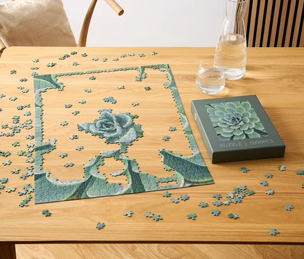 Částečně složené puzzle »Sukulenty« leží na dřevěném stole vedle krabice s motivem sukulentu, sklenice a karafy s vodou.