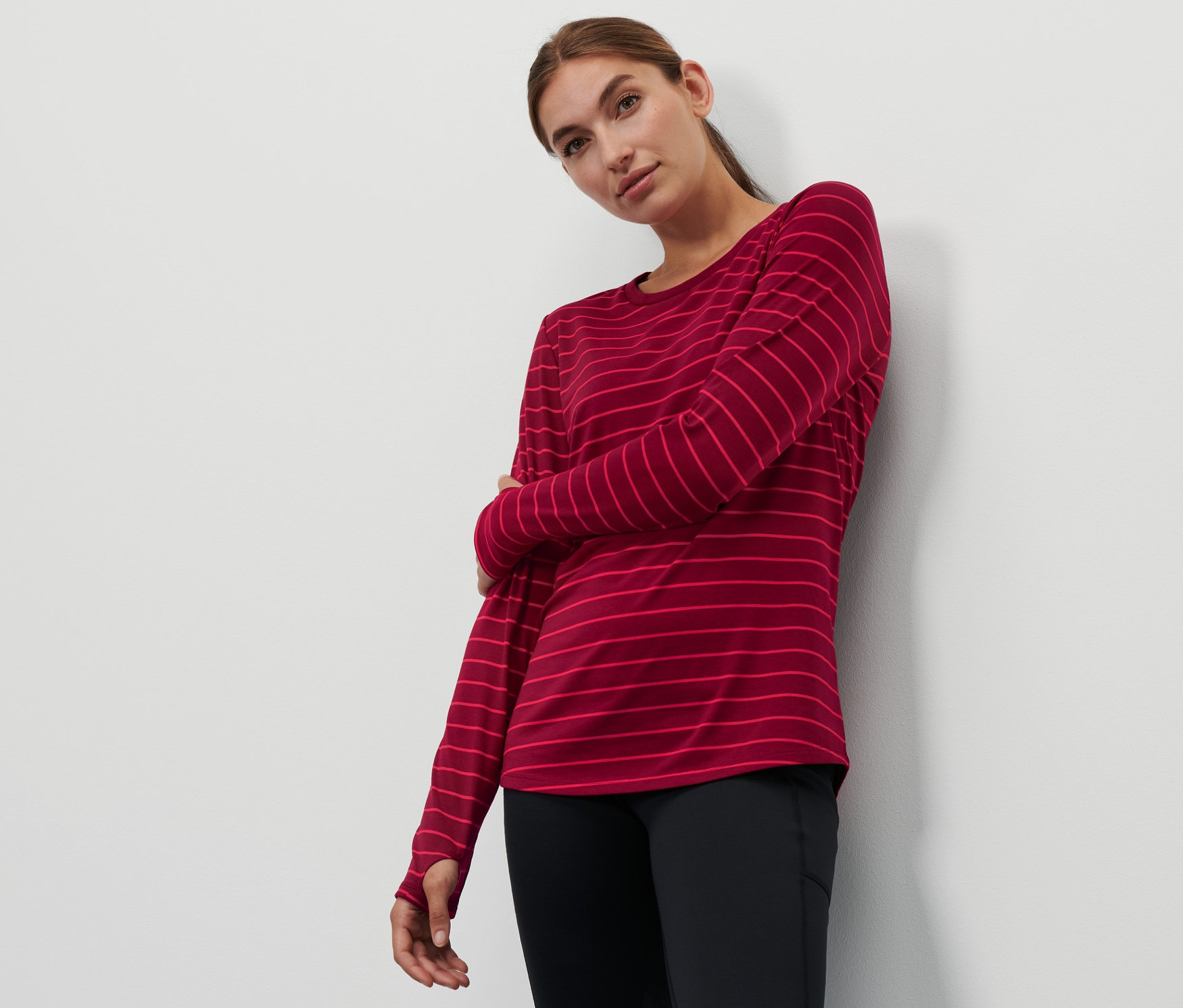 Žena pózuje v bordó pruhovaném sportovním triku s dlouhým rukávem a černých legínách.