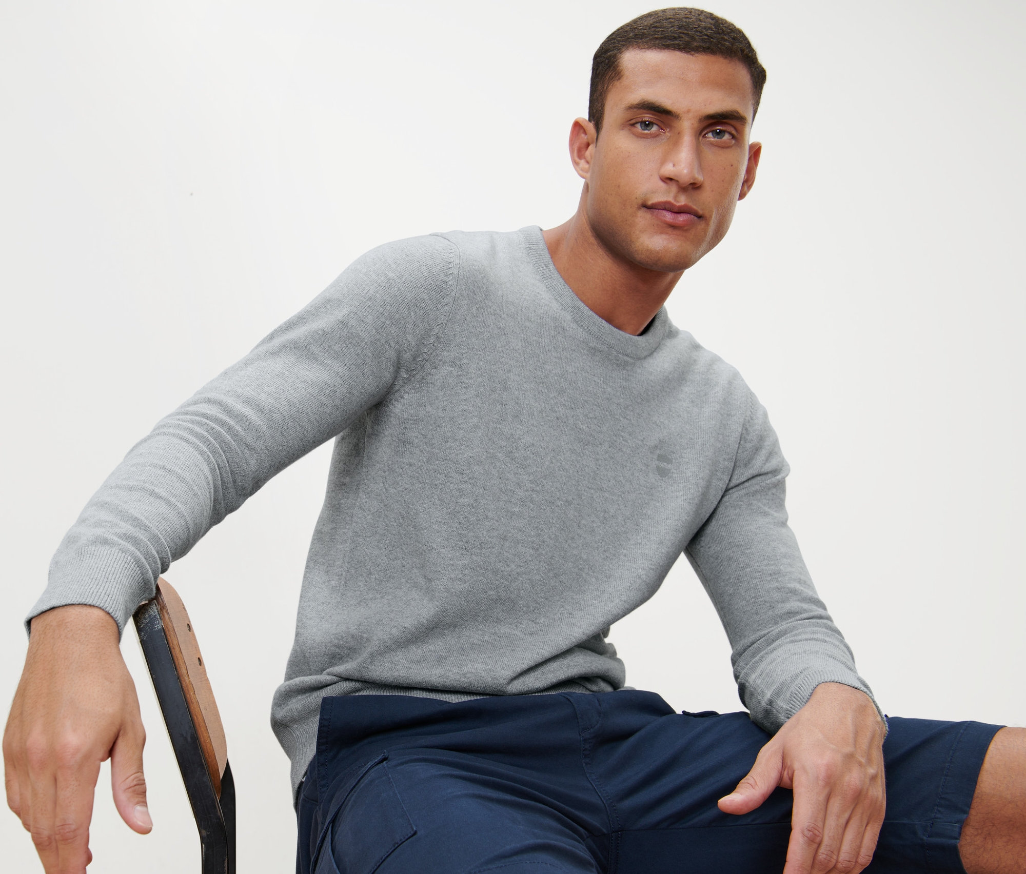 Muž sedí na židli a má na sobě šedý svetr s kulatým výstřihem.