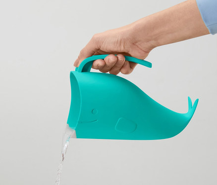 Ruka drží modrozelenou nádobku na mytí dětských vlasů ve tvaru velryby, z které teče voda.