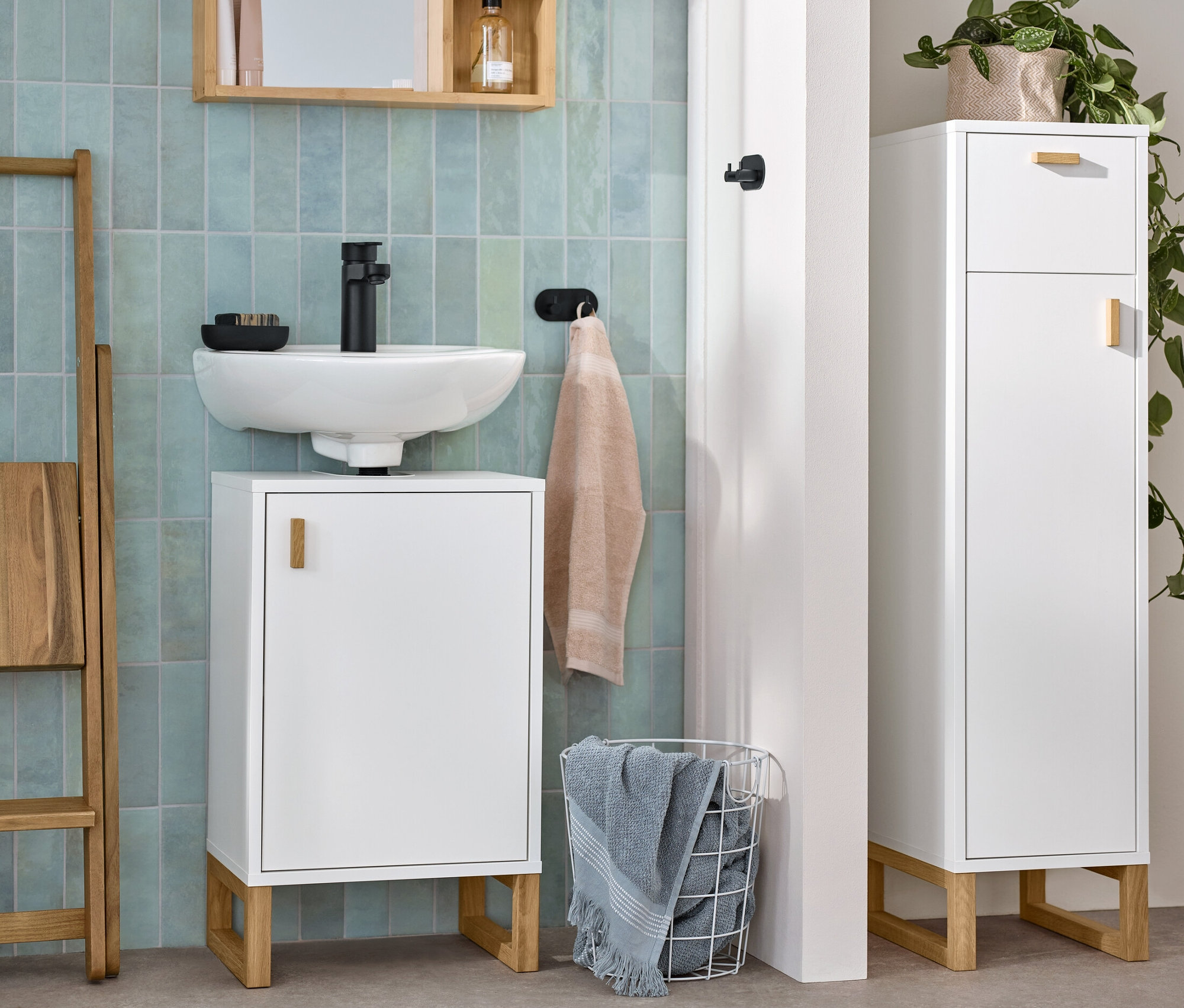 Bílá koupelna s umyvadlem, malá skříňka pod umyvadlo s dvířky, úzká odkládací koupelnová skříňka se zásuvkou a dvířky a zrcadlo s poličkami.