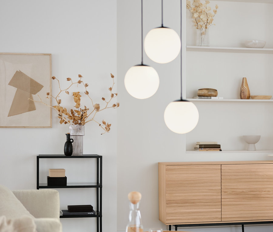 Obývací pokoj s kulatým konferenčním stolkem, komodou Finnsbo s lamelovým vzhledem a designovým konzolovým stolkem se skleněnými policemi. Nad nimi visí závěsné svítidlo se skleněnými koulemi.